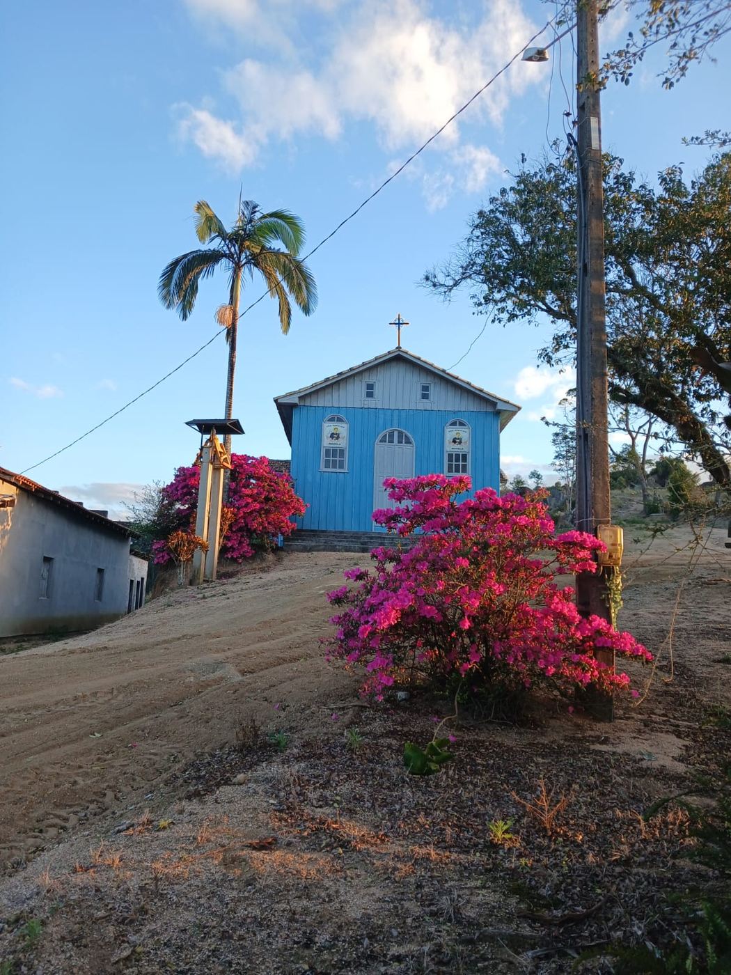 A capela no alto do morro, com azaleias em flor ao longo da subida de terra e uma palmeira ao lado.