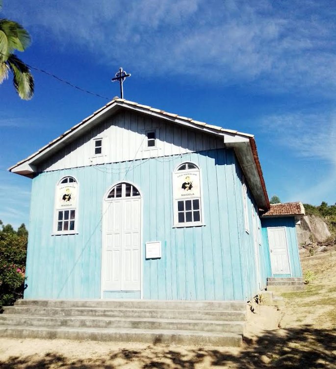 A capela vista de frente em dia de céu azul, com a cruz sobre o telhado.