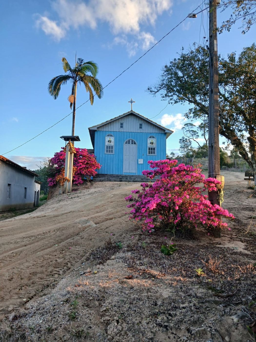 Vista aberta da estrada da comunidade, com a capela azul no alto e o sino sob a cobertura de zinco.
