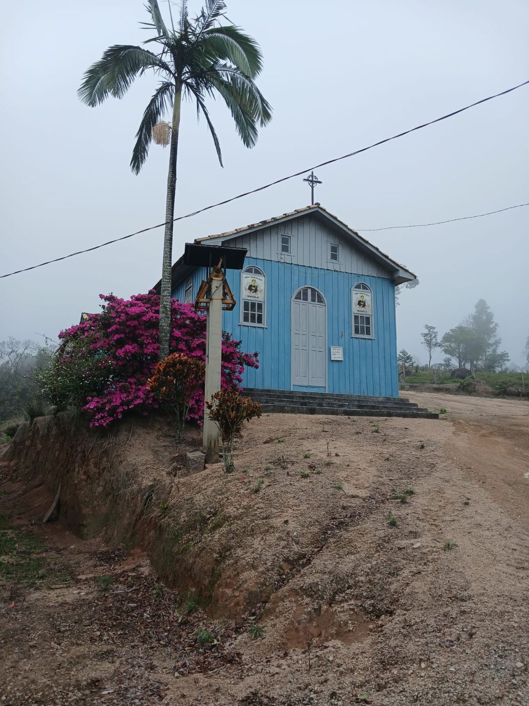 A capela em manhã de neblina, com o sino em primeiro plano e as azaleias floridas na barranca.